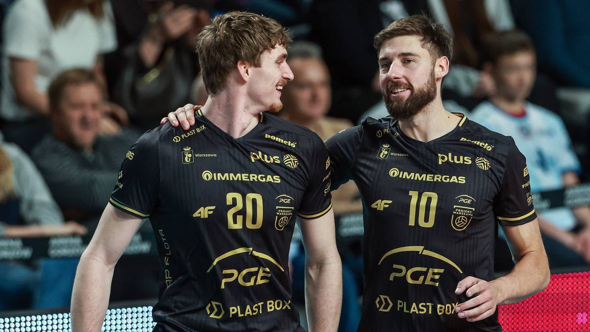 Dw&oacute;ch siatkarzy w czarnych strojach sportowych z numerami 20 i 10 na plecach, uśmiechających się do siebie na tle trybun z kibicami.