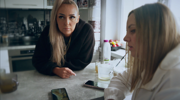 Dwie kobiety przy kuchennym blacie, jedna z nich patrzy w stronę obiektywu, druga skupiona na telefonie.