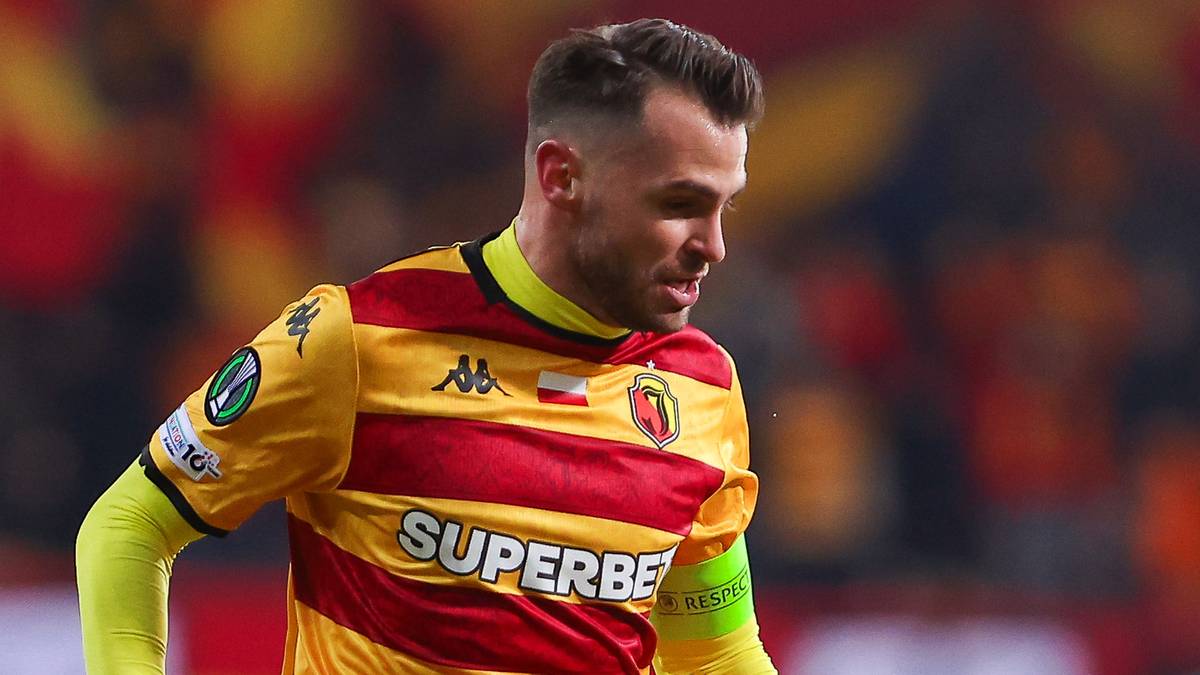 Piłkarz Jagiellonii Białystok z piłką na boisku, w tle inni zawodnicy w fioletowych strojach