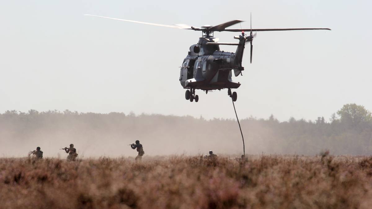 Helikopter wojskowy z opuszczonymi żołnierzami na linie, lądujący nad suchym terenem.