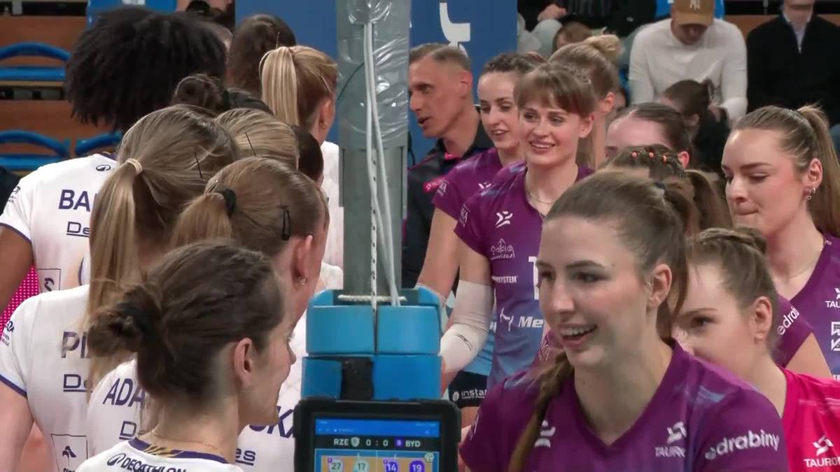 Grupa uśmiechniętych siatkarek w strojach sportowych, otoczonych przez kibic&oacute;w i personel, po meczu siatk&oacute;wki.