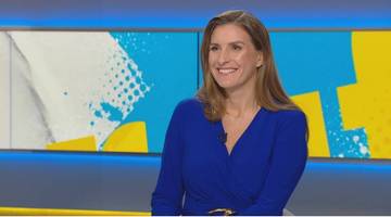 Kobieta w niebieskiej sukience siedzi na tle studia telewizyjnego z uśmiechem na twarzy.