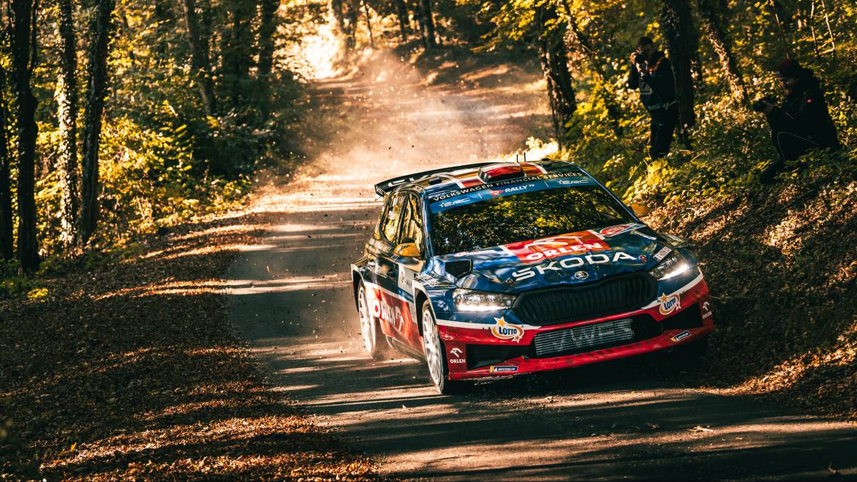 Samochód rajdowy Skoda Fabia R5 na luźnej nawierzchni leśnej drogi, unoszący kurz. Widoczne fragmenty drzew i liści.