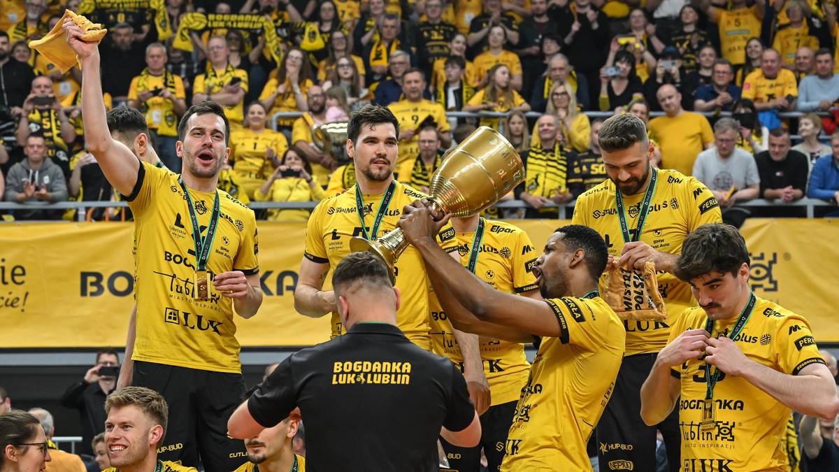 Drużyna siatkarzy w żółtych koszulkach świętuje zwycięstwo, jeden z zawodników unosi złoty puchar.