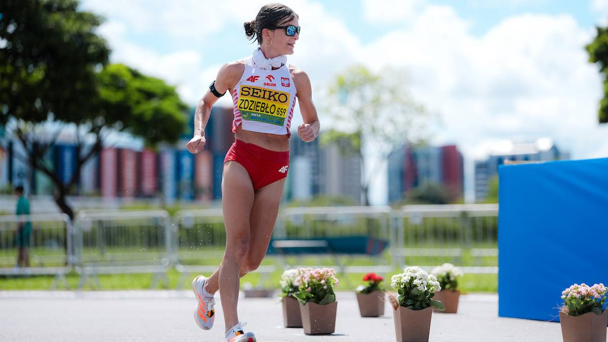 Zawodniczka w stroju sportowym biegnie po trasie, w tle budynki i zieleń.