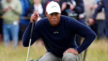 Tiger Woods klęczy na polu golfowym, mierząc uderzenie kijem golfowym, z piłką golfową przed sobą. Za nim zgromadzeni są widzowie.