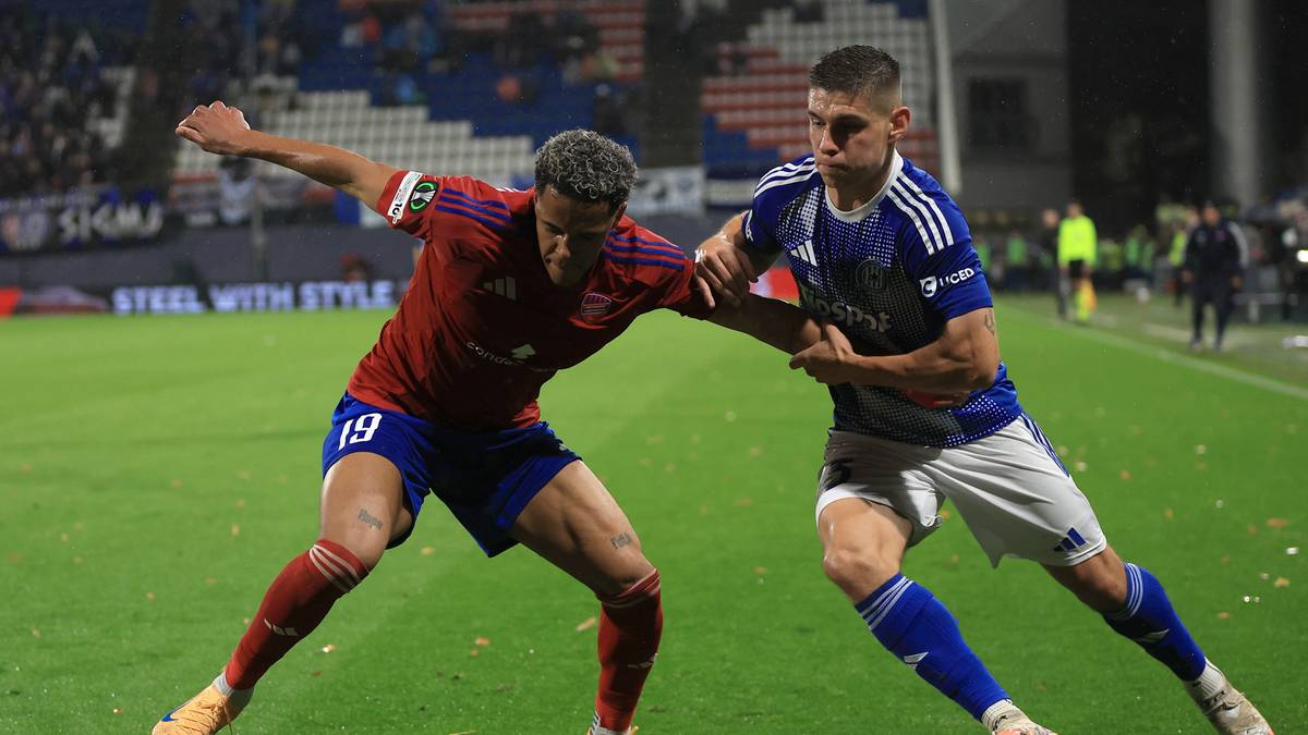 Dwóch piłkarzy walczy o piłkę na boisku. Zawodnik w czerwonej koszulce z numerem 19 i zawodnik w niebieskiej koszulce. Trawa stadionu i trybuny w tle.