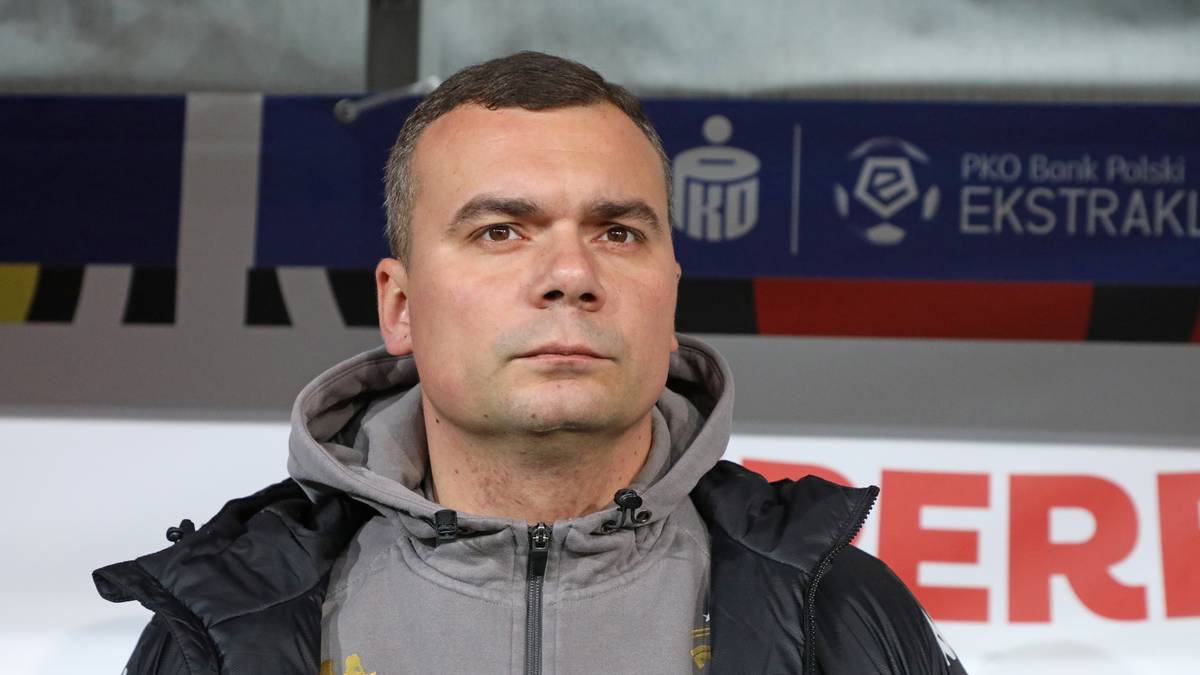 Portret mężczyzny w średnim wieku, ubranego w sportową kurtkę z kapturem. Tło stanowi baner z logo Ekstraklasy.