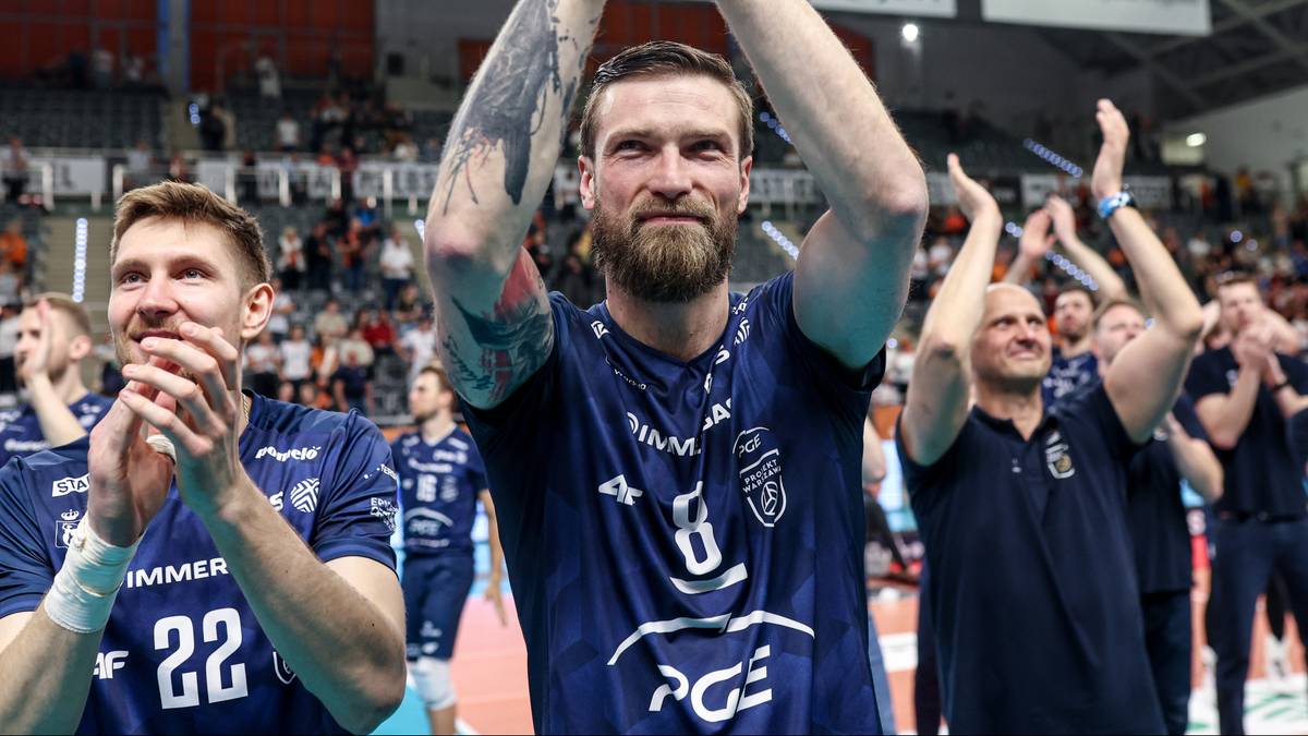 Siatkarz z brodą w niebieskiej koszulce unosi ręce w geście radości, w tle inni zawodnicy w podobnych strojach.