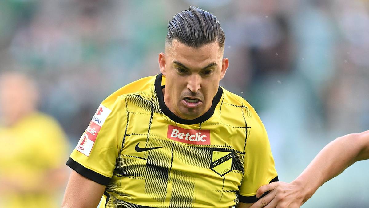 Piłkarz w żółtej koszulce z czarnymi pasami, na jego koszulce widoczne logo Betclic. Ma intensywną minę i zmierza do przodu.