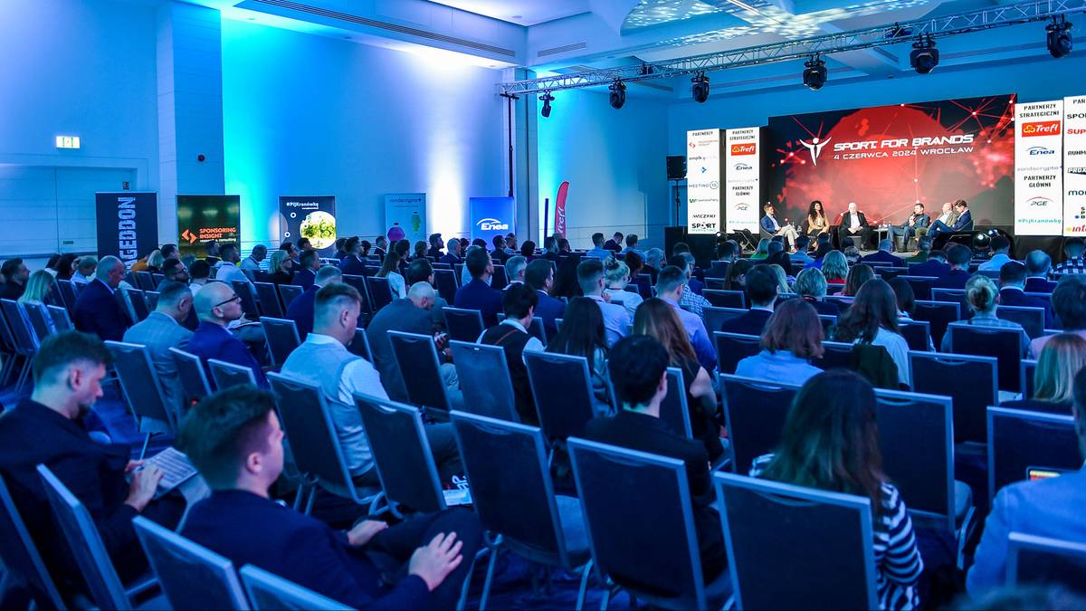 Szeroka sala konferencyjna oświetlona niebieskim światłem, z rzędem krzeseł skierowanych w stronę dużej sceny. Na scenie znajduje się ekran z logo "SPORT FOR BRANDS 2025" oraz panel dyskusyjny z kilkoma osobami. Na ekranie wyświetlane są również logotypy sponsorów. Widownia siedzi w rzędach, słuchając prelegentów.