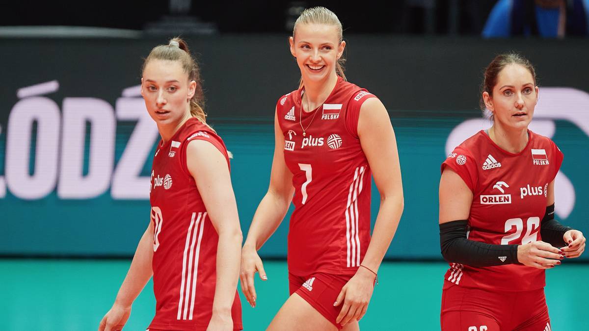 Trzy zawodniczki siatk&oacute;wki w czerwonych strojach sportowych, z numerami na koszulkach, stoją na boisku podczas meczu.