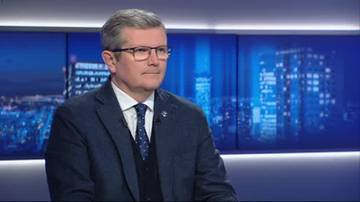 Mężczyzna w garniturze i okularach siedzi na tle ekran&oacute;w z nocnym widokiem miasta.