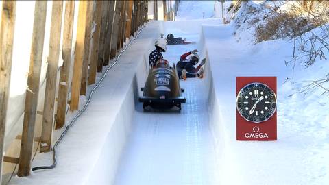 Trzech zawodnik&oacute;w w bobsleju zjeżdża po ośnieżonym torze, obok stoi baner z logo Omega.