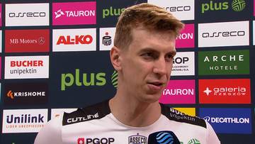 Artur Szalpuk, siatkarz, w białej koszulce z logo drużyny, na tle kolorowych logotyp&oacute;w sponsor&oacute;w.