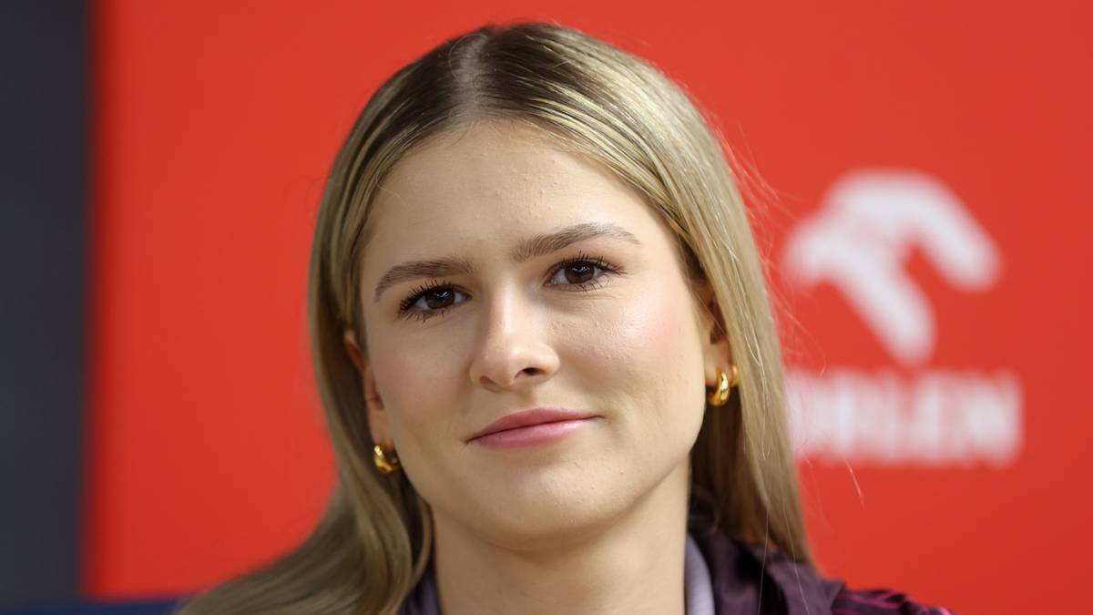 Zbliżenie na twarz młodej kobiety o jasnych włosach, uśmiechającej się lekko. W tle widać czerwony baner z białym logo.