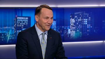 Radosław Sikorski w ciemnym garniturze i krawacie podczas wywiadu w studio telewizyjnym na tle grafiki z widokiem miasta.