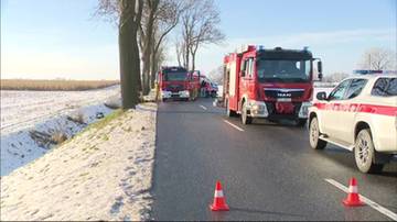 Samochód ratowniczy i wóz strażacki stoją na poboczu drogi w zimowy dzień. Widać fragmenty zaśnieżonego pola i drzew. Samochód ratowniczy ma biało-czerwone oznaczenia.