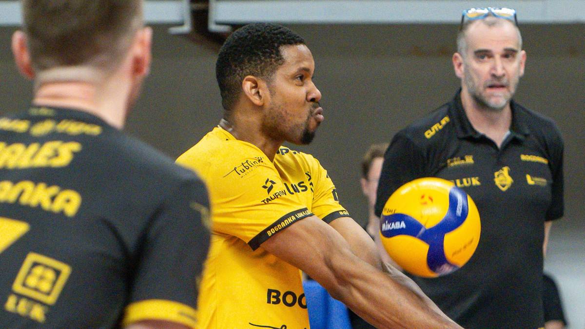 Trzech mężczyzn w strojach sportowych. W centrum, zawodnik w żółtej koszulce, zbliża się do zagrywki, patrząc przed siebie. Po jego prawej stronie, trener w czarnej koszulce monitoruje mecz, z piłką siatkową w rękach. Po lewej, widoczna jest część sylwetki innego zawodnika.