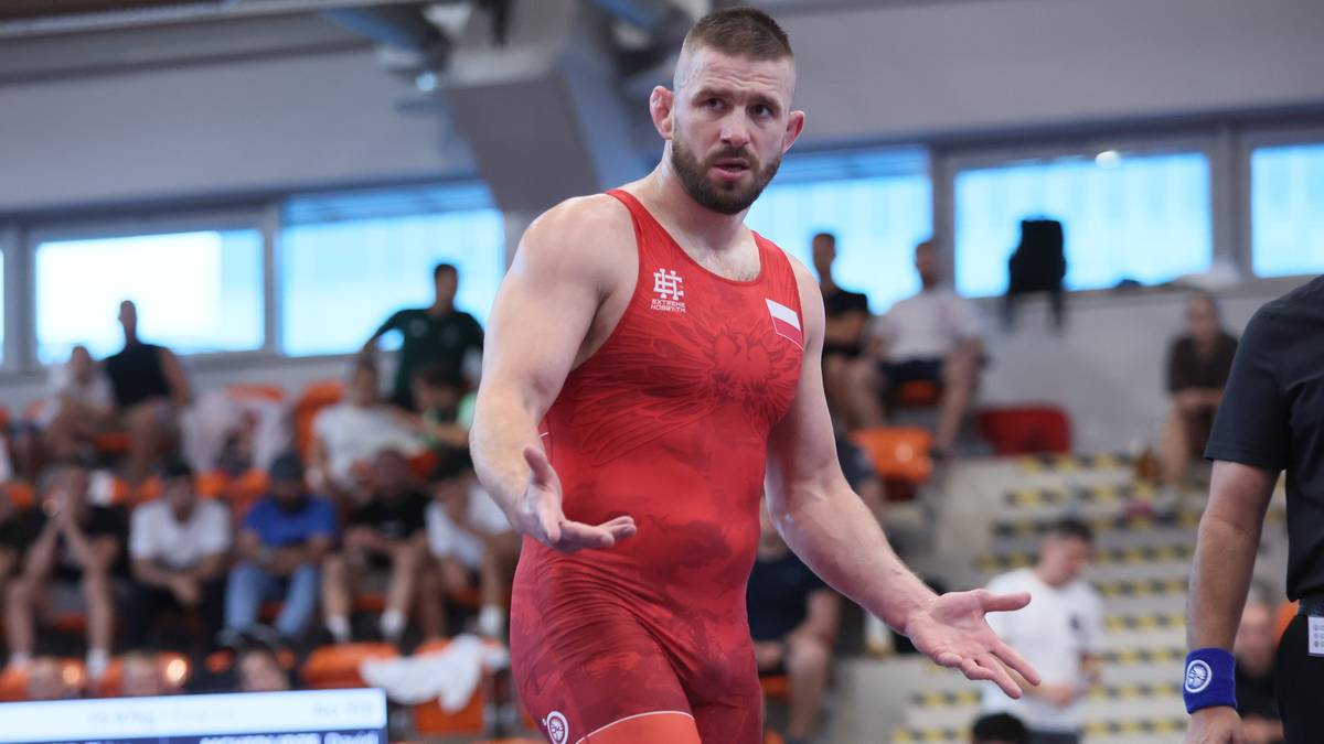 Zapaśnik w czerwonym stroju sportowym z uniesionymi dłońmi, wyrażający frustrację lub rozczarowanie.