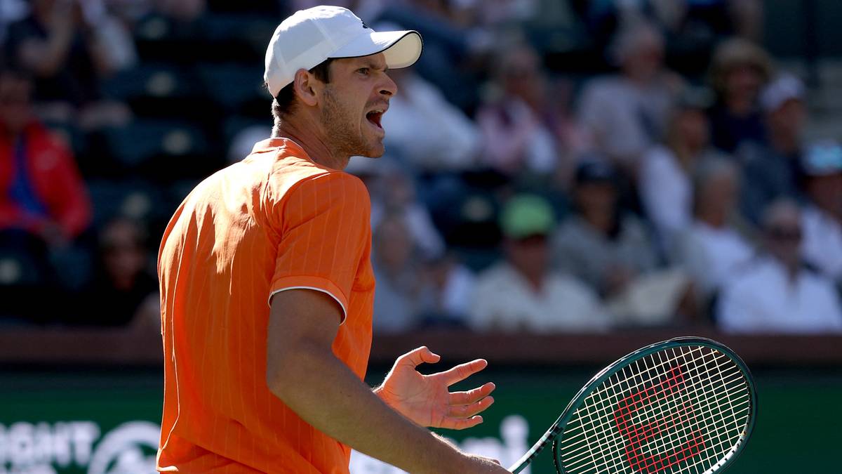 Tenisista w pomarańczowej koszulce i białej czapce z daszkiem, trzymający rakietę tenisową, w trakcie meczu.