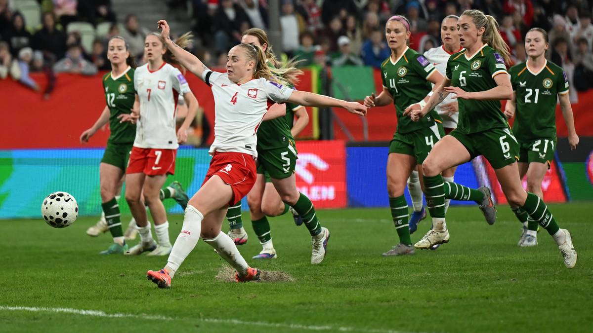 Piłkarka w białej koszulce z czerwonymi elementami drybluje z piłką na zielonej murawie, otoczona przez zawodniczki w zielonych strojach.