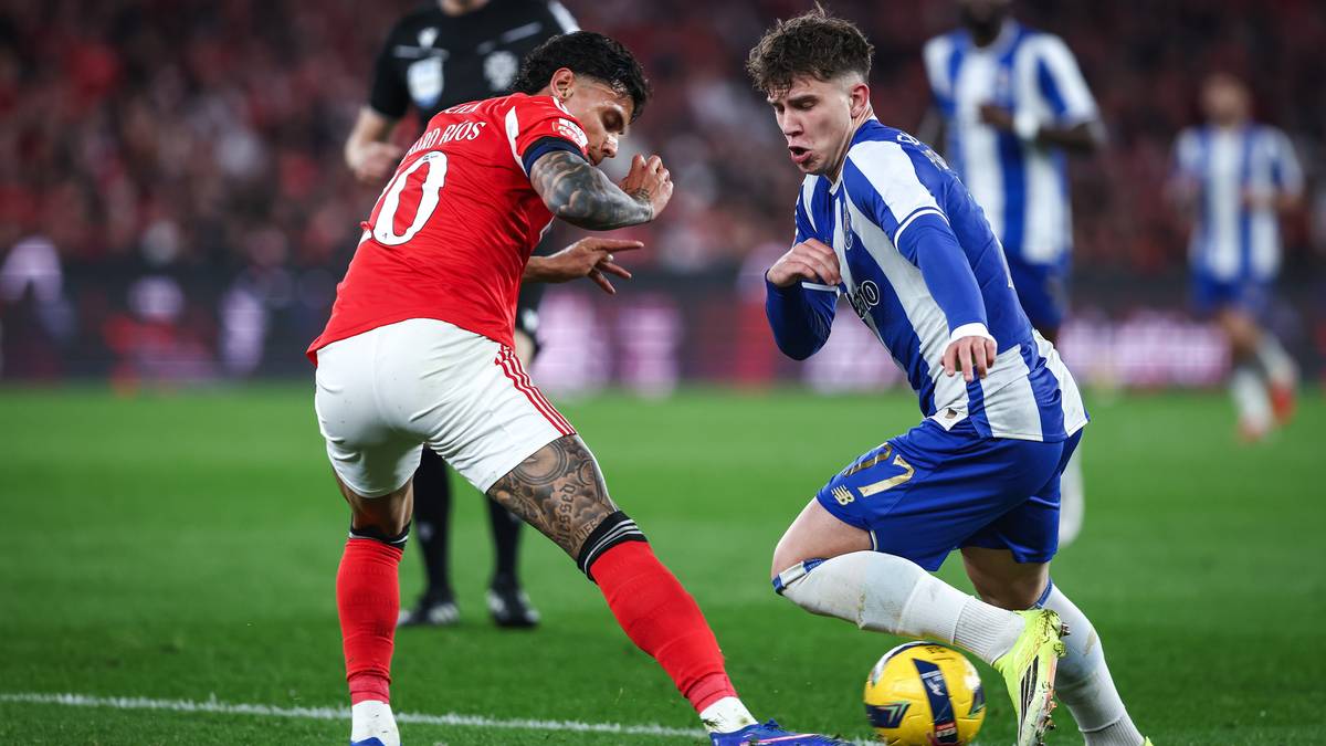 Dw&oacute;ch piłkarzy w trakcie meczu piłki nożnej, jeden w czerwonej koszulce, drugi w niebiesko-białej, walczą o piłkę na zielonej murawie stadionu.