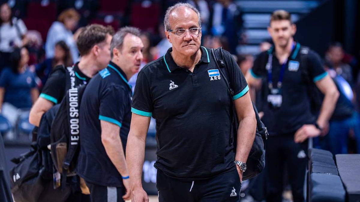 Trener koszykówki w średnim wieku, w okularach, w czarnej koszulce polo, z uwagą obserwuje boisko. Obok niego idzie inna osoba w niebieskiej koszulce z napisem "SPORT". W tle widać innych graczy i publiczność.