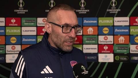 Marek Papszun, trener piłkarski, w okularach i kurtce sportowej, przemawia do mikrofonu na tle ścianki z logo Ligi Konferencji Europy i sponsor&oacute;w.