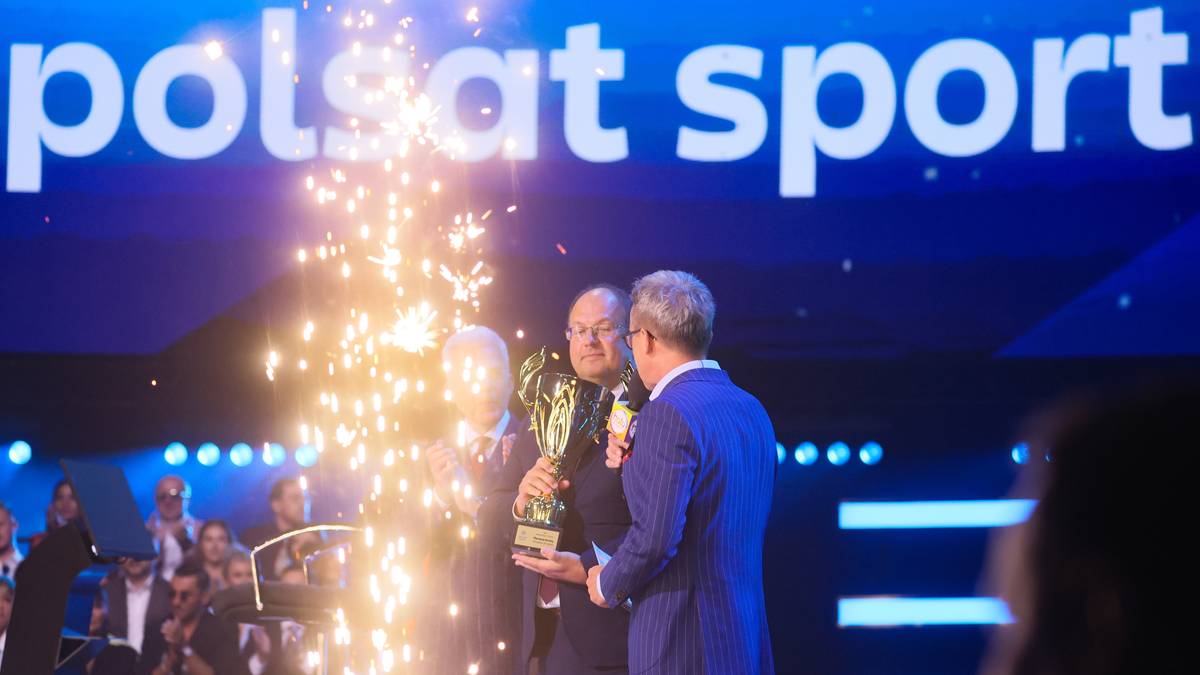 Dwóch mężczyzn na scenie. Jeden wręcza drugiemu puchar, podczas gdy z tyłu lecą fajerwerki. Na ekranie w tle widnieje napis "polsat sport".