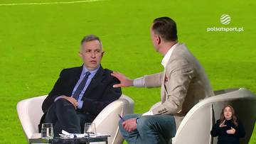 Dw&oacute;ch mężczyzn rozmawia w fotelach na tle boiska piłkarskiego. Jeden z nich, w ciemnym garniturze, siedzi, drugi, w jasnej marynarce, gestykuluje. W prawym dolnym rogu widoczna jest kobieta migająca.