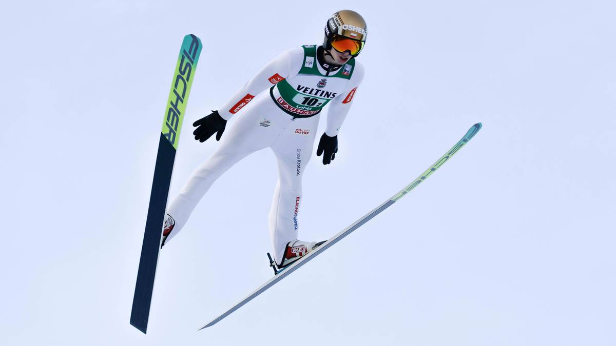 Skoczek narciarski w białym kombinezonie i kasku z goglami wykonuje skok na nartach, prezentując sylwetkę w powietrzu na tle jasnego, niebieskawego nieba.