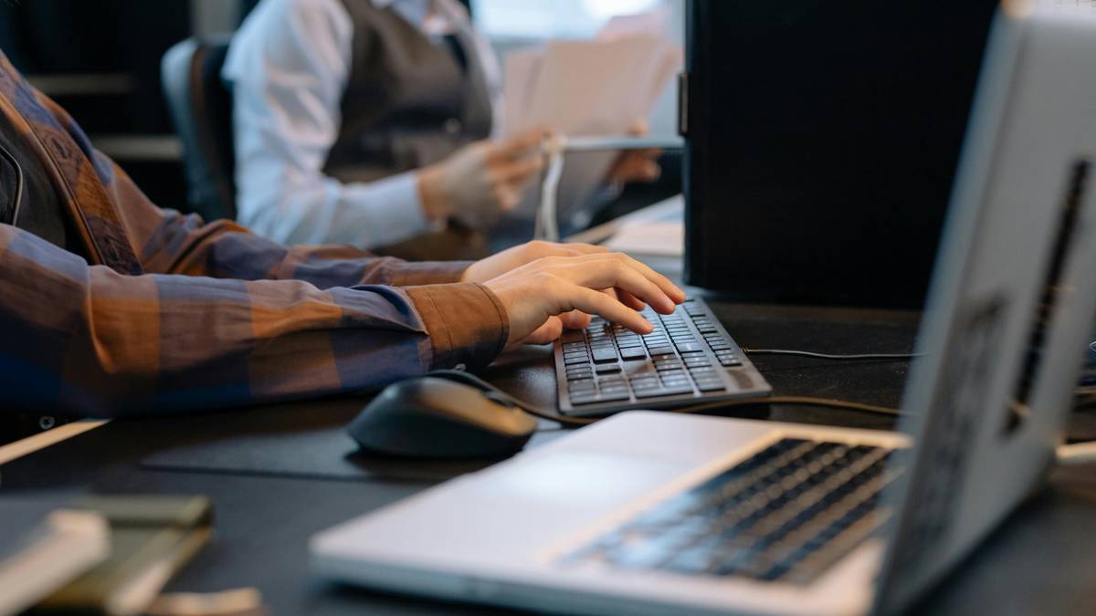 Dłonie osoby piszącej na klawiaturze laptopa, obok myszka komputerowa. W tle druga osoba przy biurku.