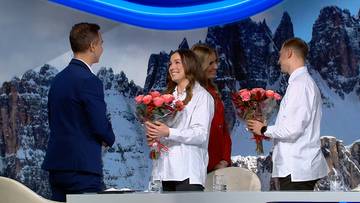 Dwie kobiety i dwóch mężczyzn w studiu telewizyjnym, na tle gór. Dwie osoby otrzymują bukiety różowych kwiatów.