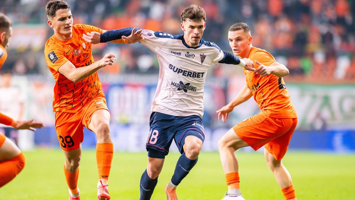 Trzech piłkarzy na boisku, dw&oacute;ch w pomarańczowych strojach walczy o piłkę z zawodnikiem w białej koszulce z numerem 8.