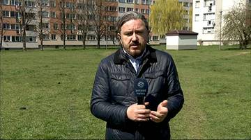 Mężczyzna w ciemnej kurtce i białej koszuli trzyma mikrofon z logo "Polsat News" i m&oacute;wi do kamery na tle blok&oacute;w mieszkalnych i zielonego trawnika.