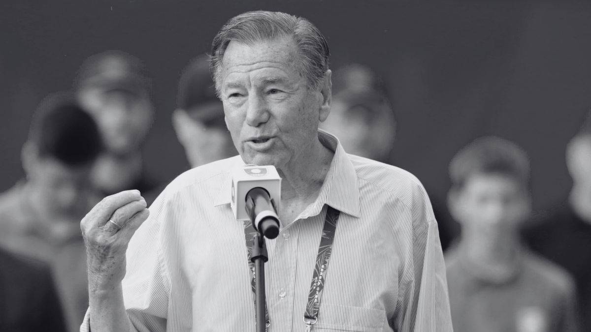 Czarno-białe zdjęcie przedstawia starszego mężczyznę, Rod Laver, mówiącego do mikrofonu, z niewyraźnymi sylwetkami ludzi w tle.