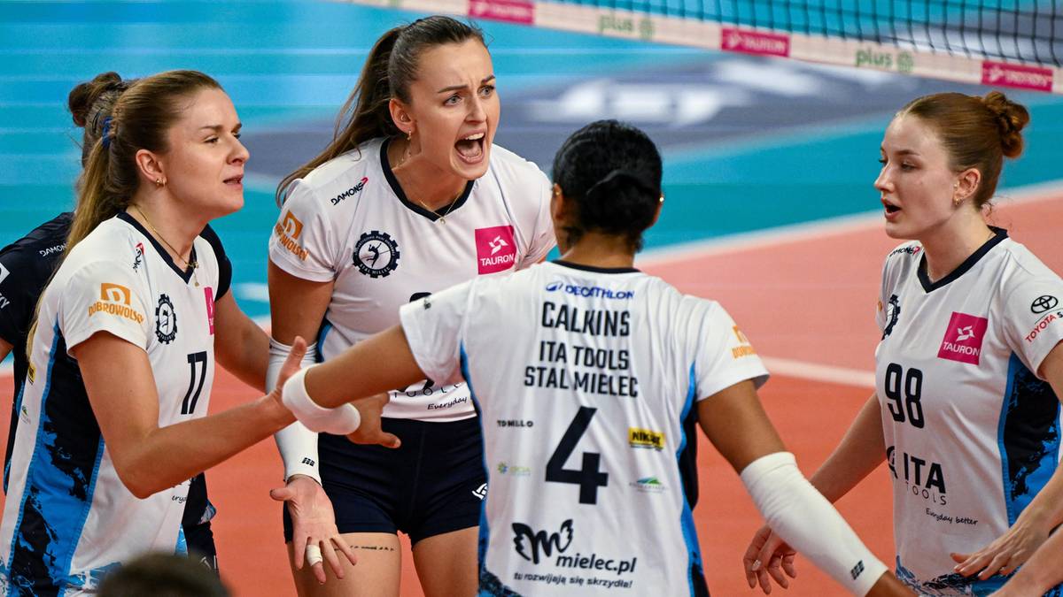 Grupa siatkarek w białych strojach z numerami na plecach celebruje zwycięstwo na boisku siatkarskim.