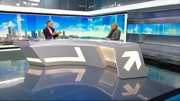 Dw&oacute;ch mężczyzn siedzi przy futurystycznym stole w studiu telewizyjnym. Jeden z nich, ubrany w jasną koszulę, gestykuluje, przemawiając. Drugi, w ciemniejszym stroju, słucha go uważnie. W tle widać panoramiczny widok miasta.