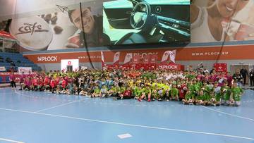 Grupa dzieci w strojach sportowych siedzi na środku boiska hali sportowej.