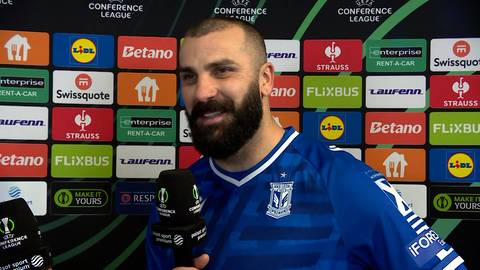 Mikael Ishak, piłkarz Lecha Poznań, w niebieskiej koszulce z herbem klubu, na tle ścianki z logotypami sponsor&oacute;w.