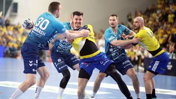 Piłkarze ręczni w akcji podczas meczu, jeden zawodnik w ż&oacute;łto-niebieskim stroju jest atakowany przez trzech graczy w niebieskich koszulkach.