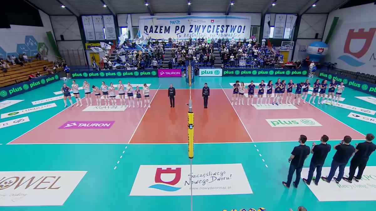 Dwie drużyny siatkarek stoją po przeciwnych stronach siatki na boisku, przed rozpoczęciem meczu. Na trybunach zgromadzeni są kibice.