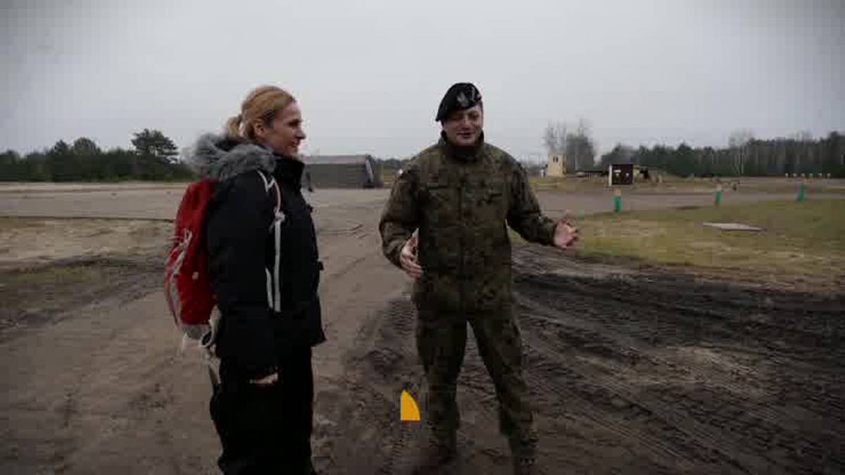 Dziennikarka Polsatu w czarnej kurtce i z czerwonym plecakiem rozmawia z żołnierzem w mundurze polowym i czarnym beretem na poligonie.