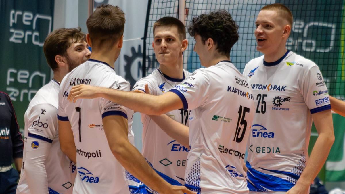 Sześciu siatkarzy w białych strojach z niebieskimi elementami, stojących w kręgu i rozmawiających.