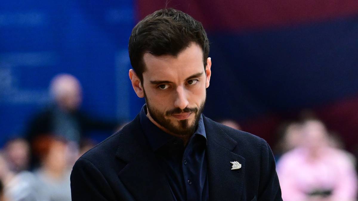 Mężczyzna w ciemnym garniturze z zaciętą miną, z dłońmi złożonymi przed sobą, na tle publiczności podczas wydarzenia sportowego.