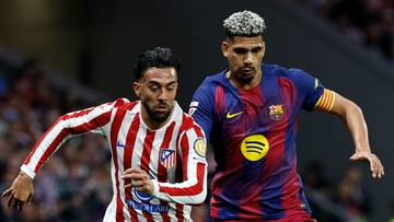 Dw&oacute;ch piłkarzy w akcji na boisku, jeden w stroju Atletico Madryt, drugi w stroju FC Barcelony, walczą o piłkę.