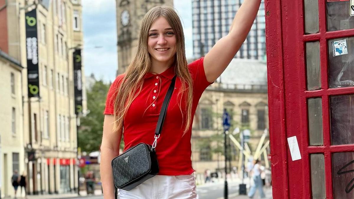 Młoda kobieta w czerwonej koszulce polo i białych szortach stoi przy czerwonej budce telefonicznej. Uśmiecha się i patrzy w stronę obiektywu. W tle widać zabudowania miejskie.