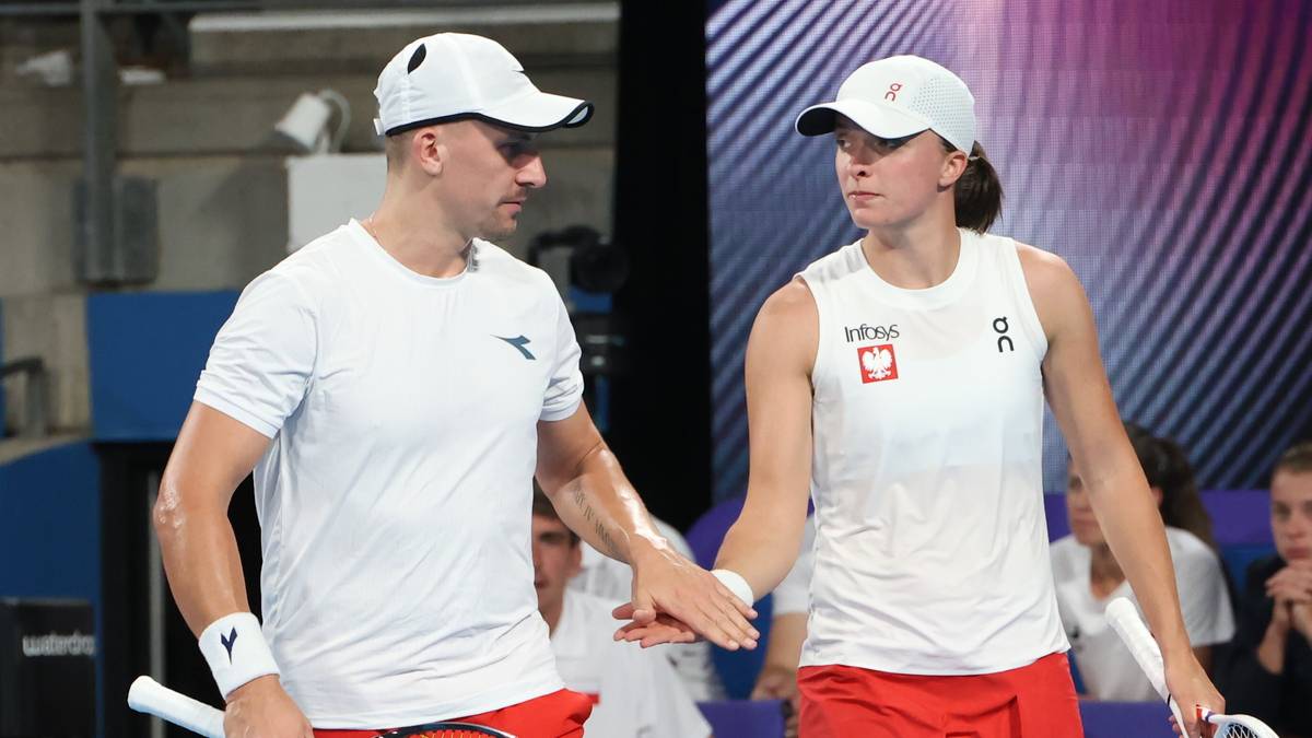 Para tenisistów w białych koszulkach i czerwonych spodenkach stoi obok siebie, patrząc w różne strony. W tle widać innych ludzi i elementy infrastruktury sportowej.