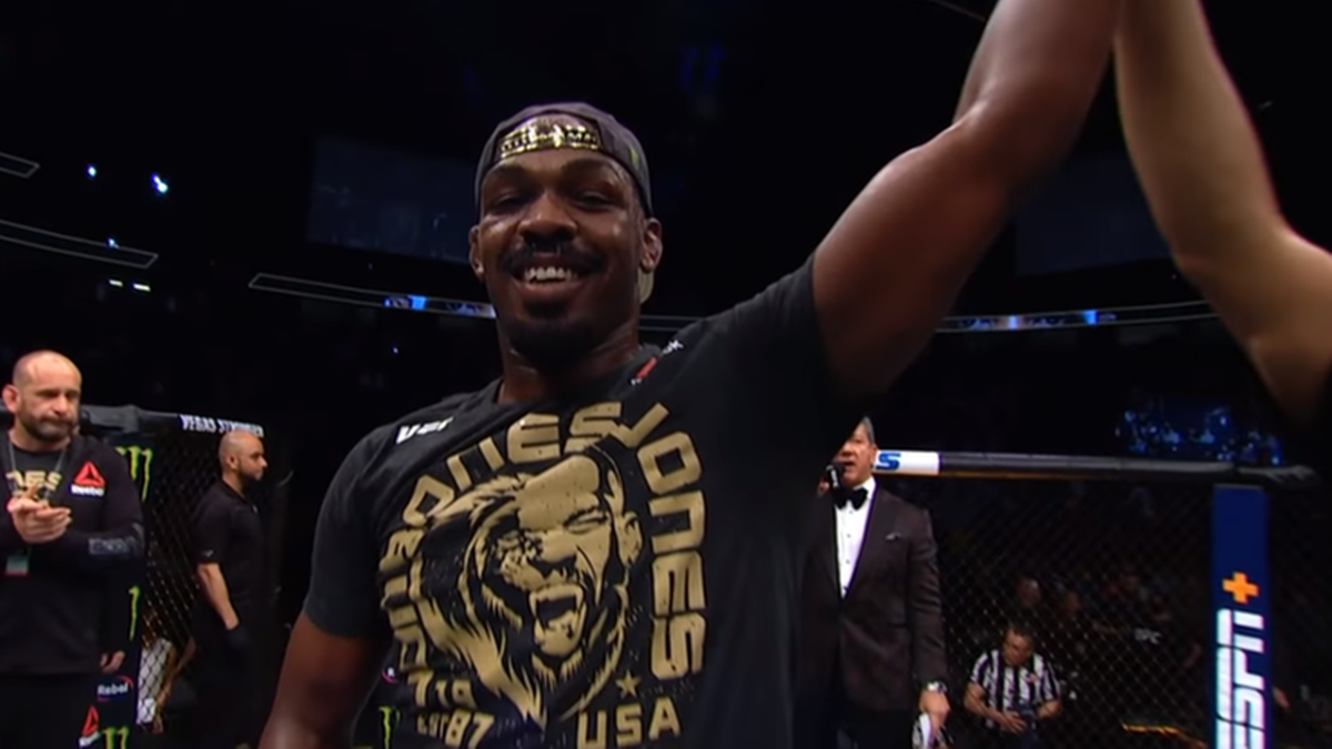 Jon Jones w czarnej koszulce z napisem "JONES" i logo, z uniesioną ręką, uśmiecha się do publiczności na gali MMA.